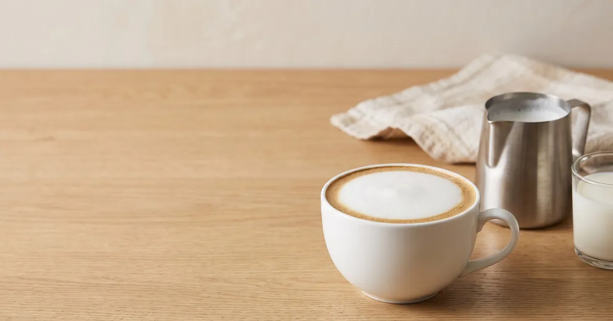 KI-generiertes Symbolbild: weite Tasse mit Milchschaum, Edelstahl-Kännchen und ein Glas Milch auf heller Arbeitsplatte